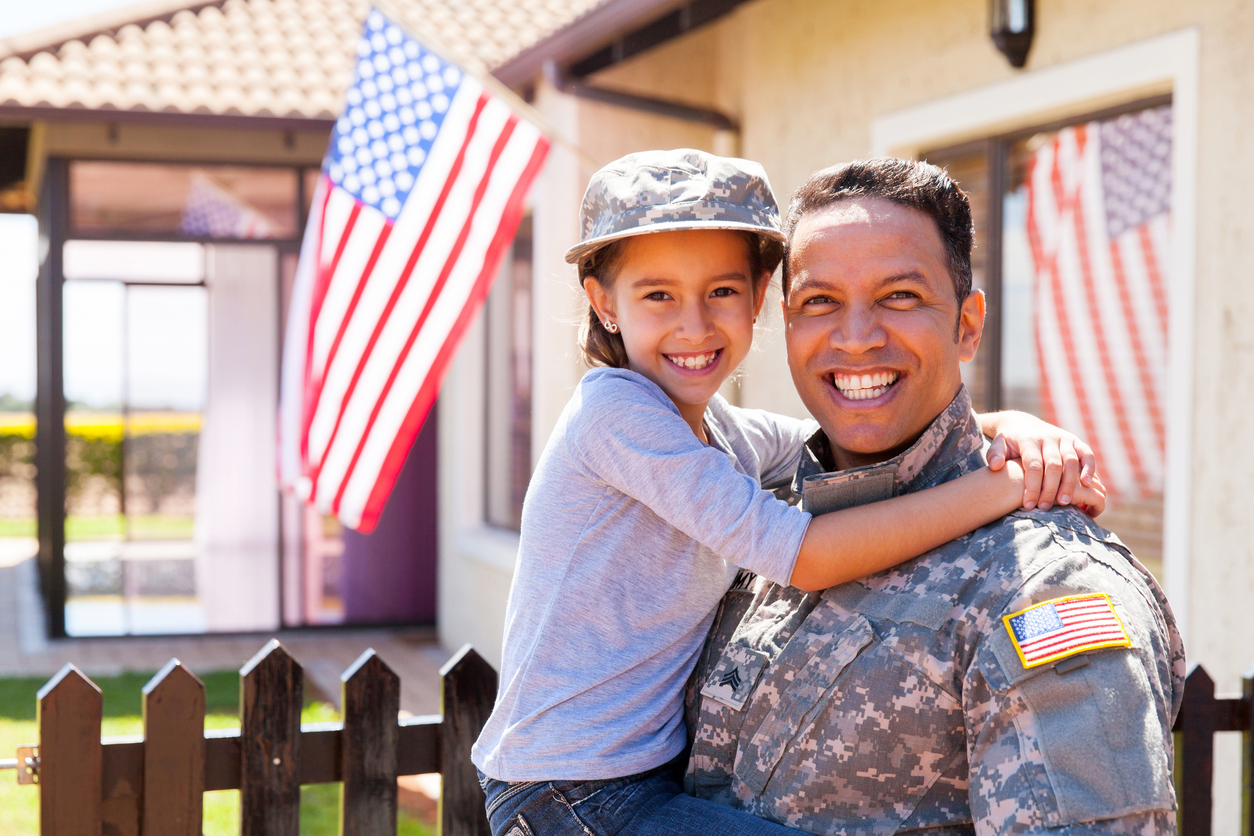 A portrait of a US Army soldier and holding his daughter outside their home; both are smiling.
